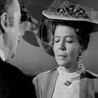 Black-and-white film still: a woman wearing a hat looks down while a man in profile faces her and speaks.