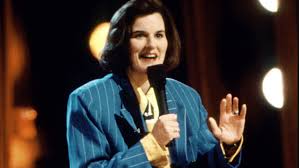 Female comedian on stage holding a microphone and waving, wearing a blue blazer.