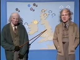 Two presenters stand before a UK weather map; one holds a pointer, pointing at weather icons like clouds and rain on the map behind them.