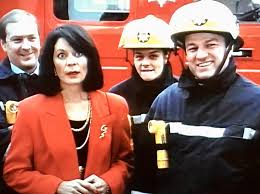 Woman in a red blazer posing with two firefighters in front of a red fire truck outside a vehicle bay.