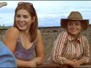 Two smiling women leaning on a railing outdoors; one in a purple tank top with sunglasses, the other in a striped shirt and wide-brim hat.