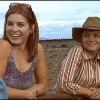 Two smiling women leaning on a railing outdoors; one in a purple tank top with sunglasses, the other in a striped shirt and wide-brim hat.