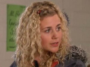 Woman with curly blonde hair looks to the side in a classroom, a green poster is on the wall behind her.