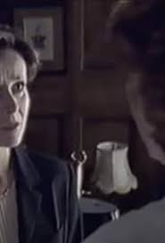 Close-up of a woman in a dark blazer speaking to a blurred figure in a dim wood-paneled room.