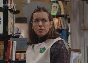 Woman wearing a white vest and glasses stands among bookshelves in a bookstore, looking toward the camera.