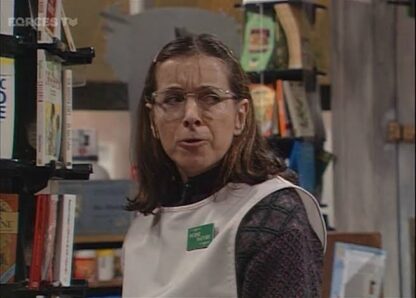 Woman wearing a white vest and glasses stands among bookshelves in a bookstore, looking toward the camera.