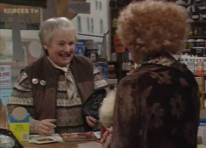 Two elderly women in a shop, a smiling cashier behind the counter greeting a customer facing away from the camera.