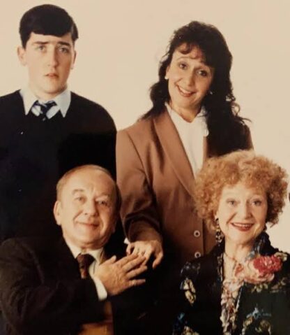 Four people pose for a family portrait: a young man in a dark sweater, a smiling woman in a brown blazer behind them, and an older couple seated at the front with the woman wearing a floral scarf.