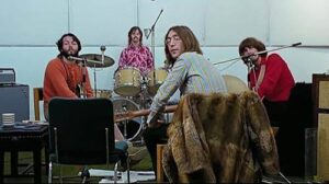 Four musicians rehearse in a studio: guitarist at center-right wearing a striped shirt, drummer in back, bassist in red on the right, and another player in orange on the left; a fur coat draped over a chair in front.