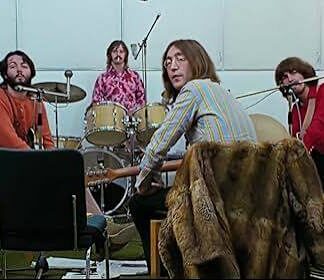 Four musicians rehearse in a studio: guitarist at center-right wearing a striped shirt, drummer in back, bassist in red on the right, and another player in orange on the left; a fur coat draped over a chair in front.