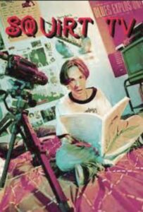 Young person sits cross-legged reading a magazine while a video camera on a tripod films in a colorful, poster-filled room.