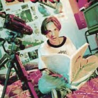 Young person sits cross-legged reading a magazine while a video camera on a tripod films in a colorful, poster-filled room.