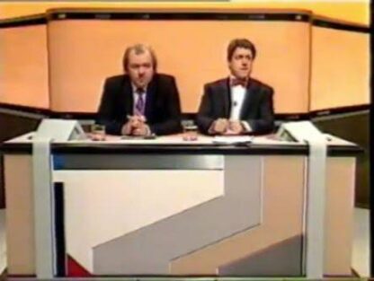 Two male news presenters sit at a modern TV desk in an orange studio set.