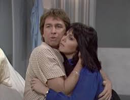 Man and woman share a close hug; the man in a beige shirt looks surprised while the woman in blue hugs him from the side.