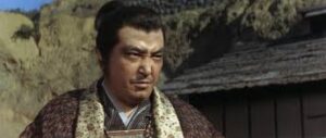 Man in traditional Japanese clothing outdoors with rocky hillside and a wooden building behind him, looking to the side.