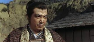 Man in traditional Japanese clothing outdoors with rocky hillside and a wooden building behind him, looking to the side.