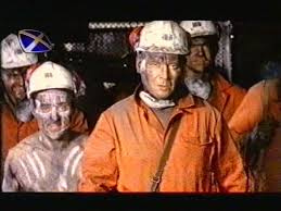 Group of miners in orange protective suits and hard hats with headlamps standing in a dark mine shaft, looking toward the camera.