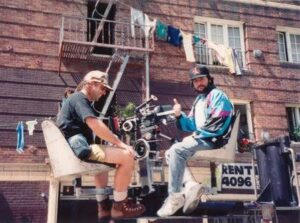 Two men work on a film shoot on a wheeled platform attached to a brick apartment building, with laundry hanging above.