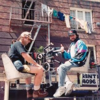 Two men work on a film shoot on a wheeled platform attached to a brick apartment building, with laundry hanging above.