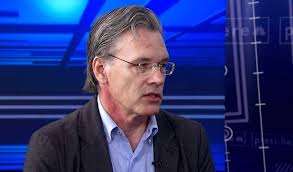Man with gray hair and glasses speaks during a TV studio interview in a blue, high-tech backdrop.